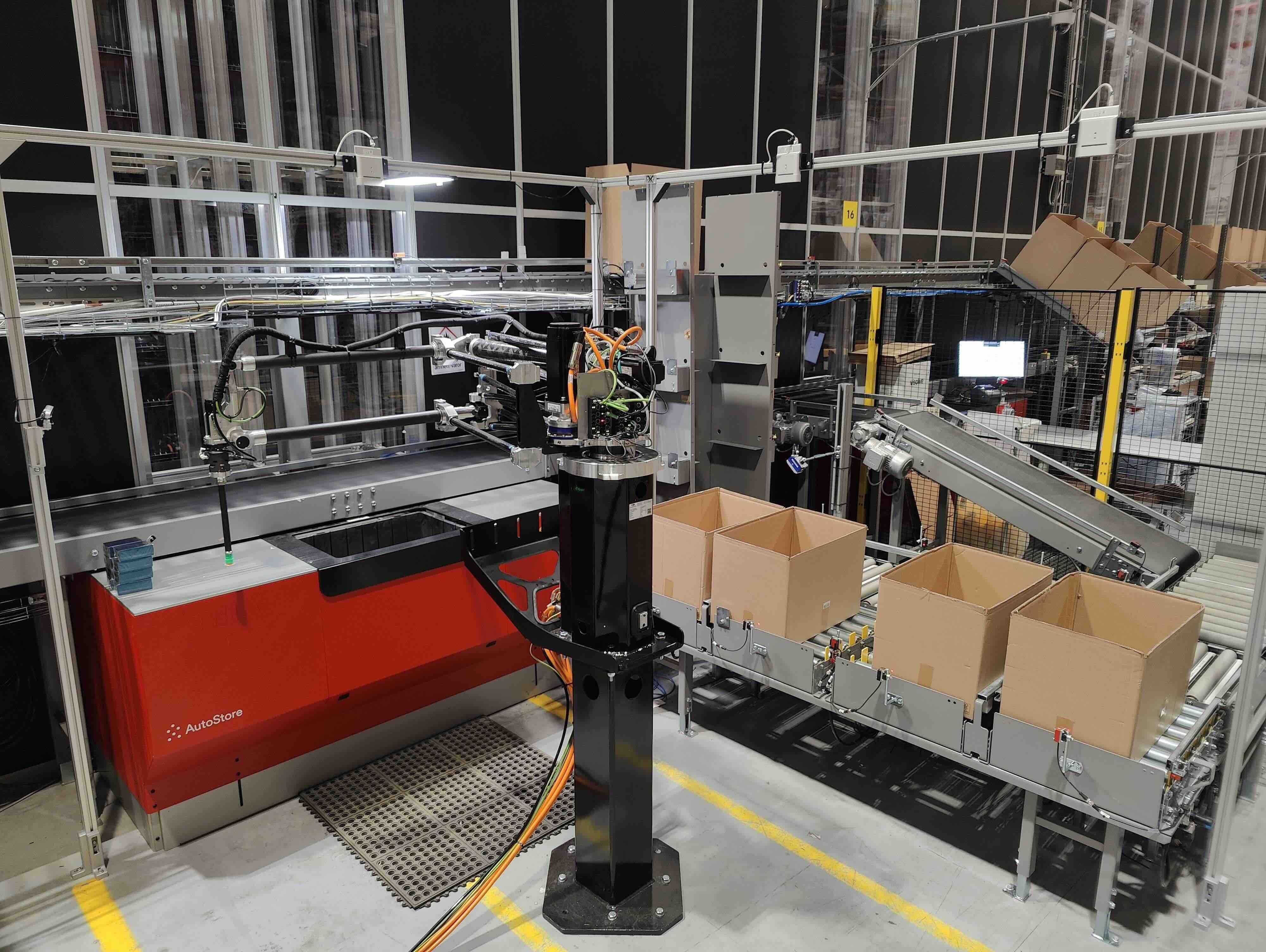 Robotic arm system in a Nowaste warehouse, sorting items into boxes on a conveyor belt, surrounded by machinery and storage.