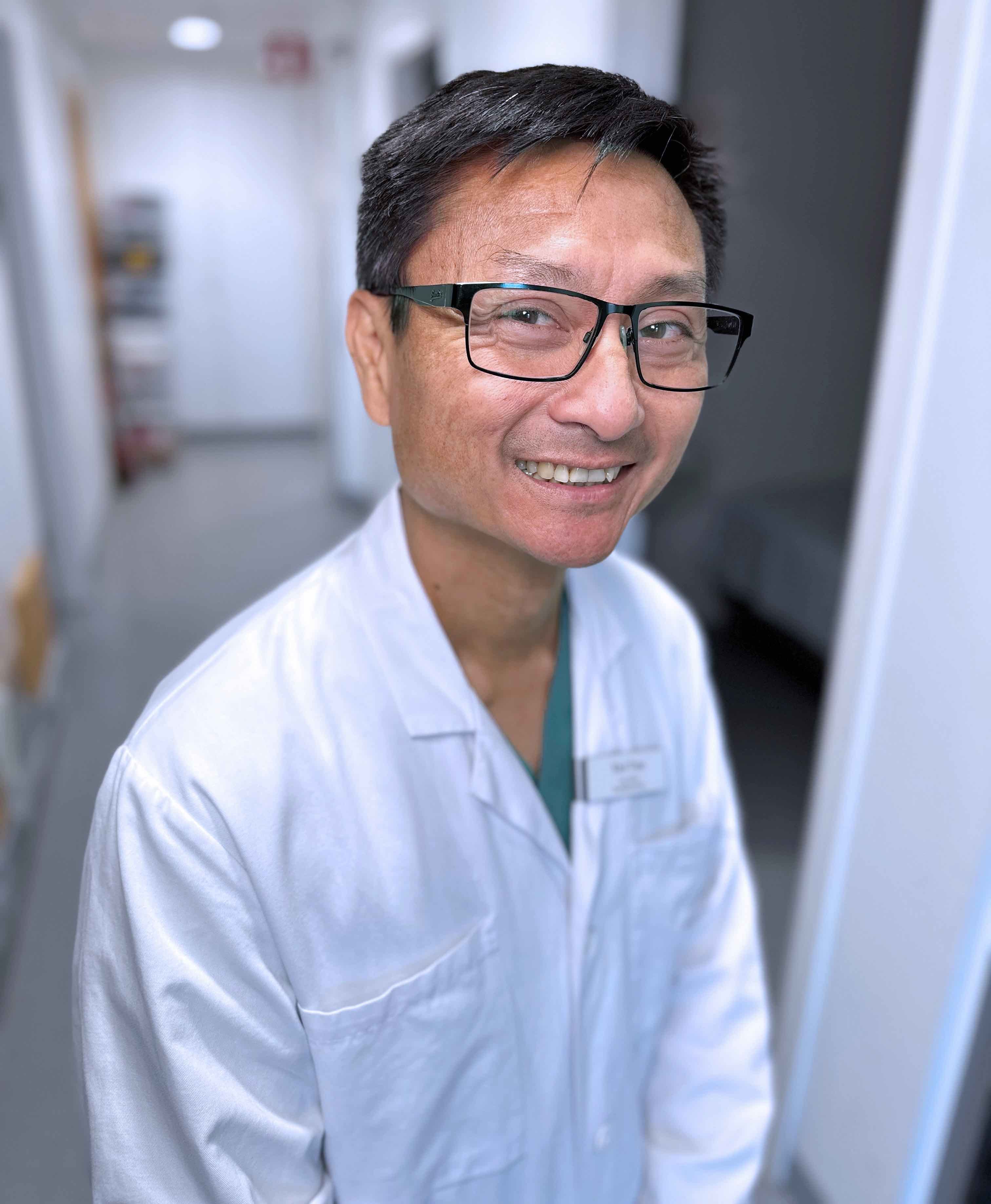 A doctor wearing glasses and a white lab coat stands in a hallway, suggesting a medical or scientific environment.