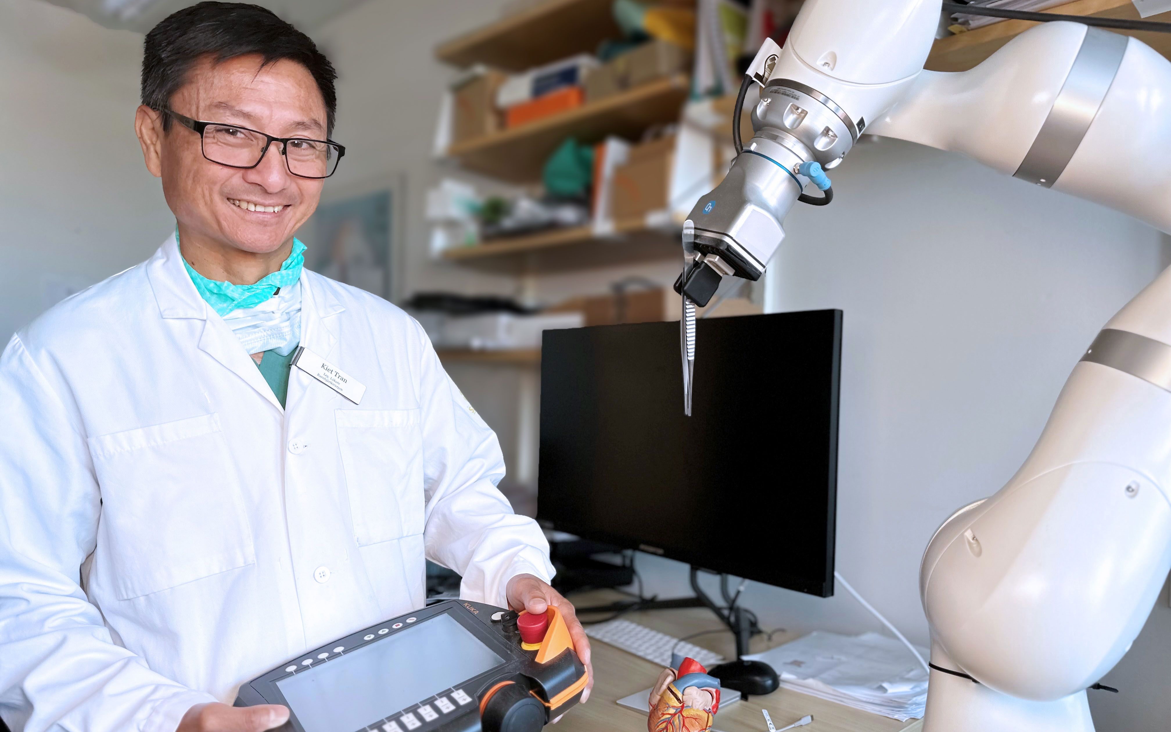 A person in a lab coat holds a control panel beside a robotic arm in a laboratory setting, with a computer and anatomical models in the background.